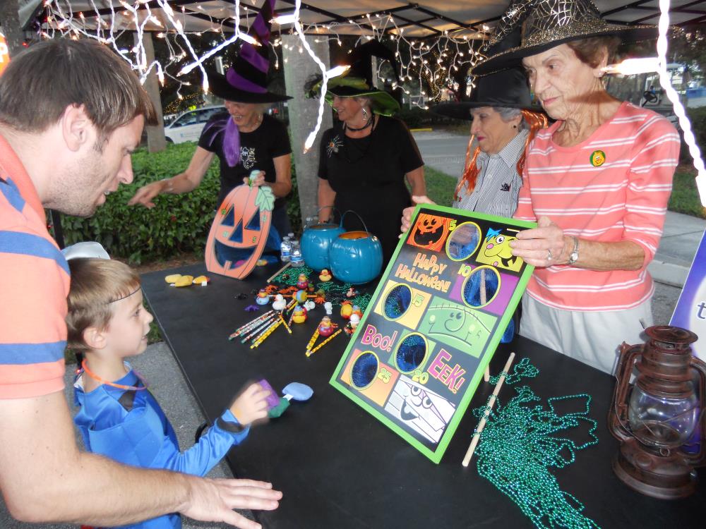 Boy tries to throw bean bags through holes in cardboard rectangle as woman in witch hat holds the cardboard